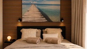 Un dormitorio con cabecera de madera, una cama grande con ropa de cama color beige, dos toallas enrolladas, una mesita de noche con una lámpara y un cuadro de un muelle sobre el agua.