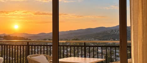Un balcón con mobiliario de mimbre y una mesa, con vistas panorámicas a montañas y una masa de agua al atardecer.