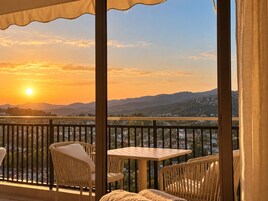 Un balcón con mobiliario de mimbre y una mesa, con vistas panorámicas a montañas y una masa de agua al atardecer.