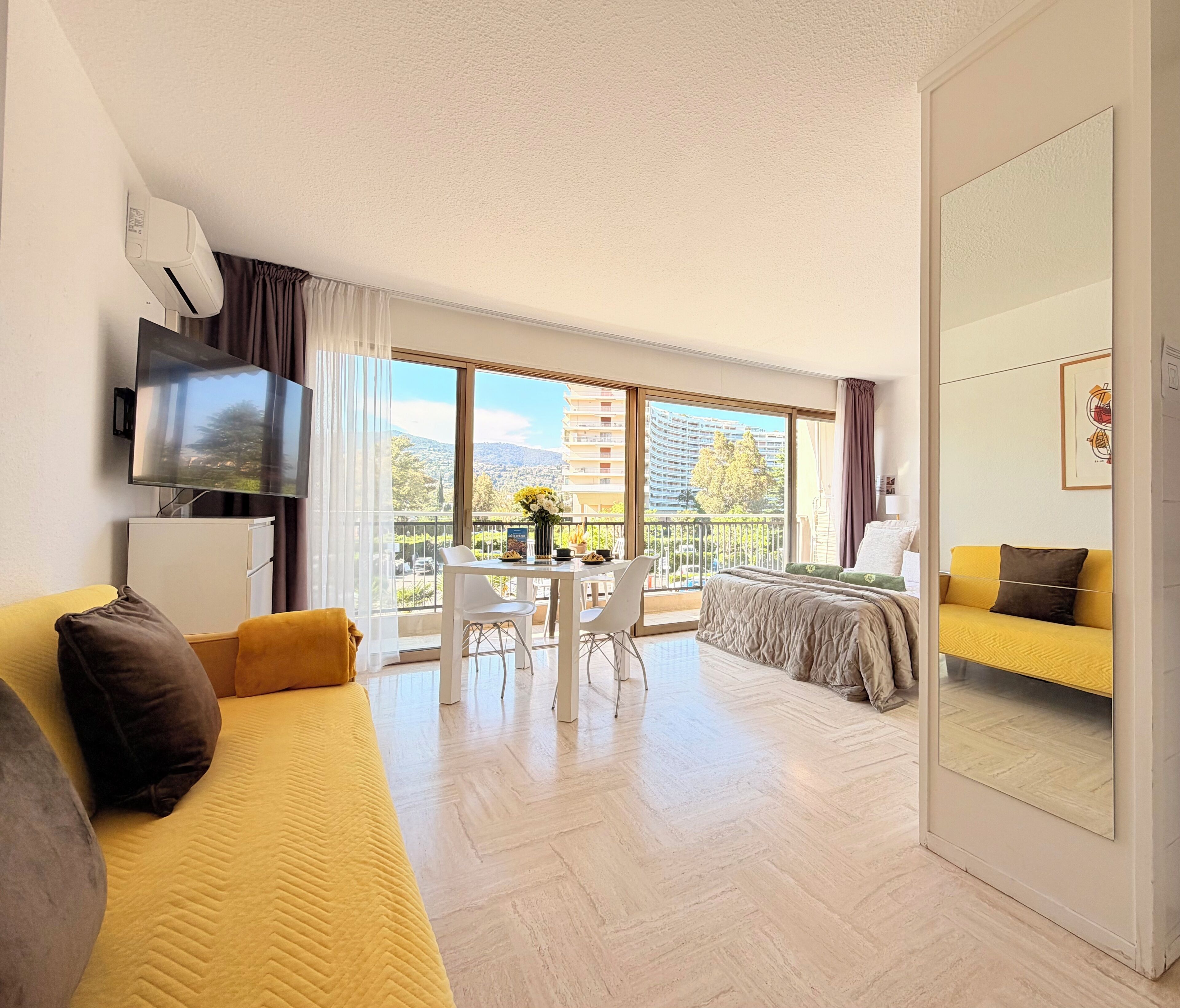 Una habitación de hotel moderna con balcón, televisión de pantalla plana, zona de comedor y cama con ropa de cama amarilla.
