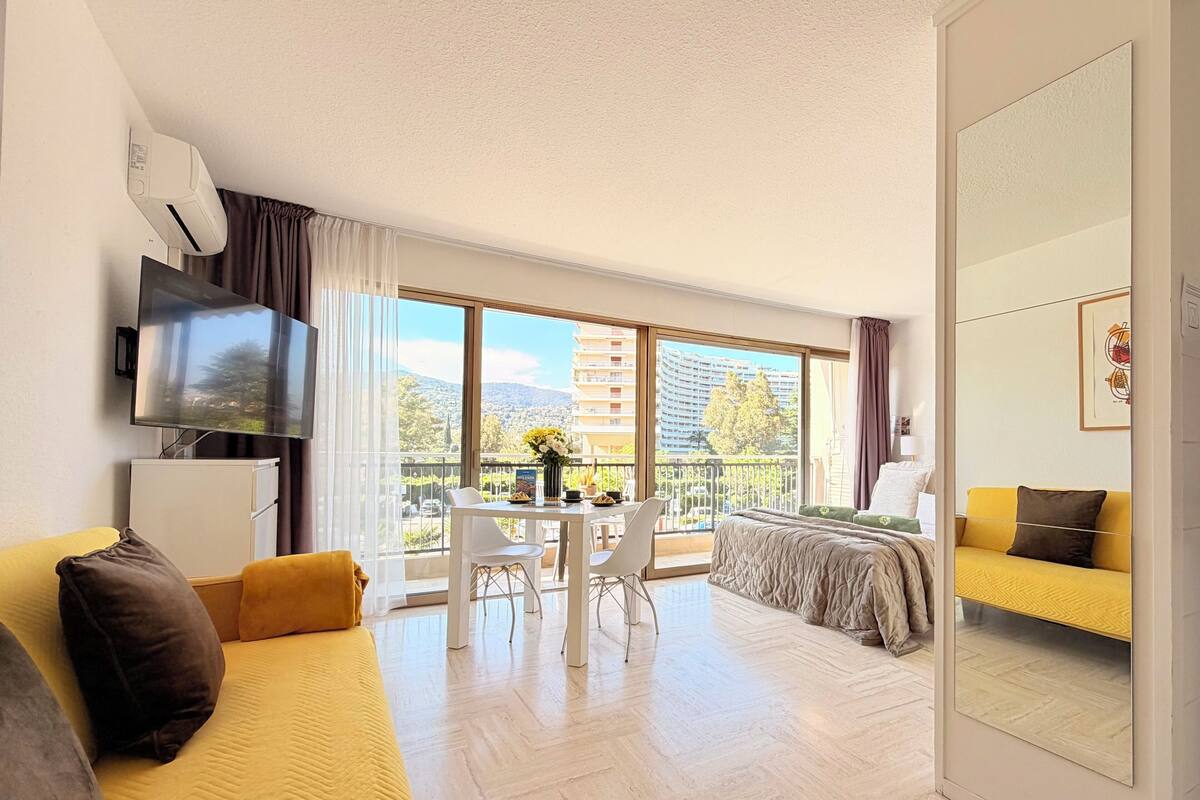 A modern hotel room with a balcony, a flat-screen TV, a dining area, and a bed with yellow bedding.