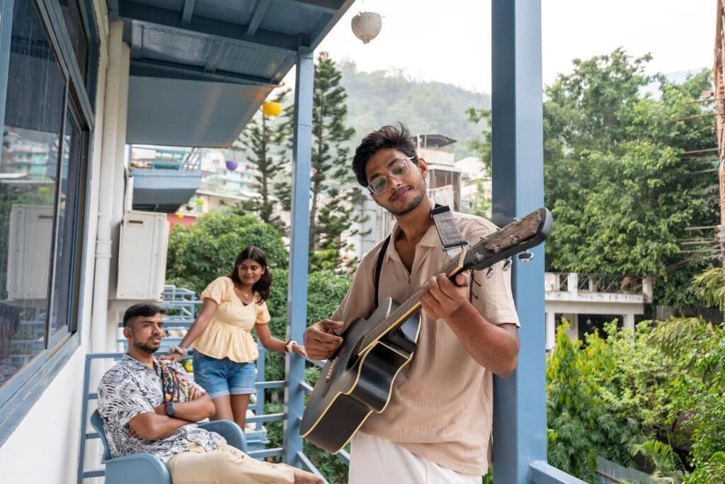 Un homme joue de la guitare sur un balcon d’où l’on peut admirer un paysage verdoyant et des bâtiments.