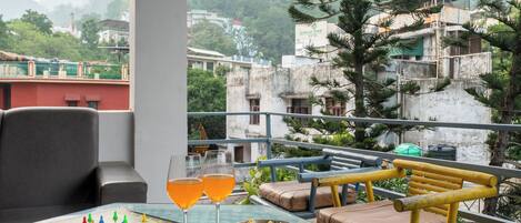 A balcony with a chessboard, two glasses of orange drink, a bowl of snacks, and a view of buildings and trees.