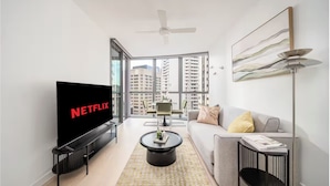 A modern living room with a large flat-screen TV, a ceiling fan, a sofa, a coffee table, and a dining area with a view of high-rise buildings. - Skyscraper Cinema - Dream a thousand city lights (Brisbane City)