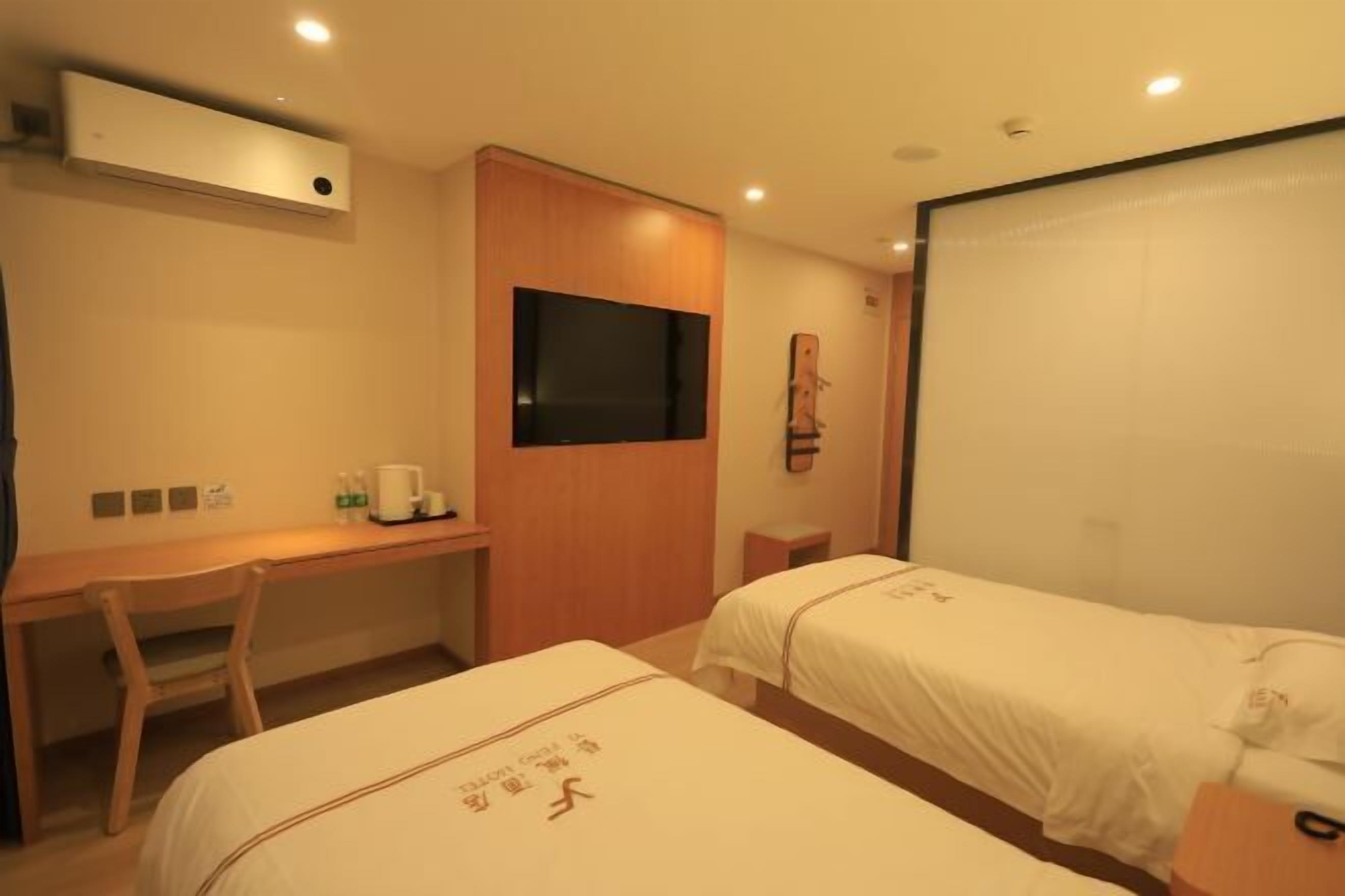 A hotel room with two single beds, a desk with a chair, a television, and a wall-mounted air conditioner.