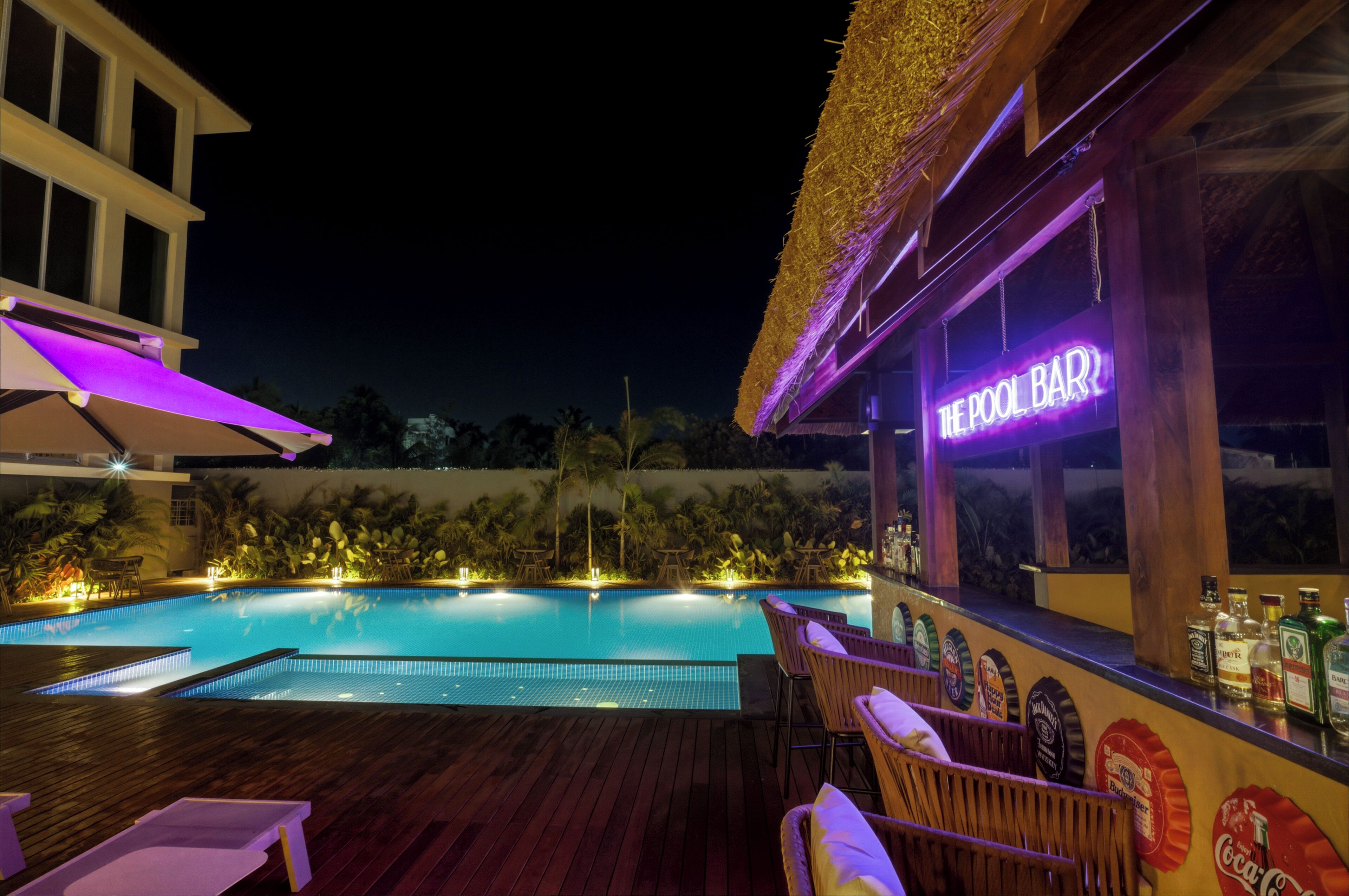 A poolside bar with a neon sign, wooden deck, lounge chairs, and a pool.