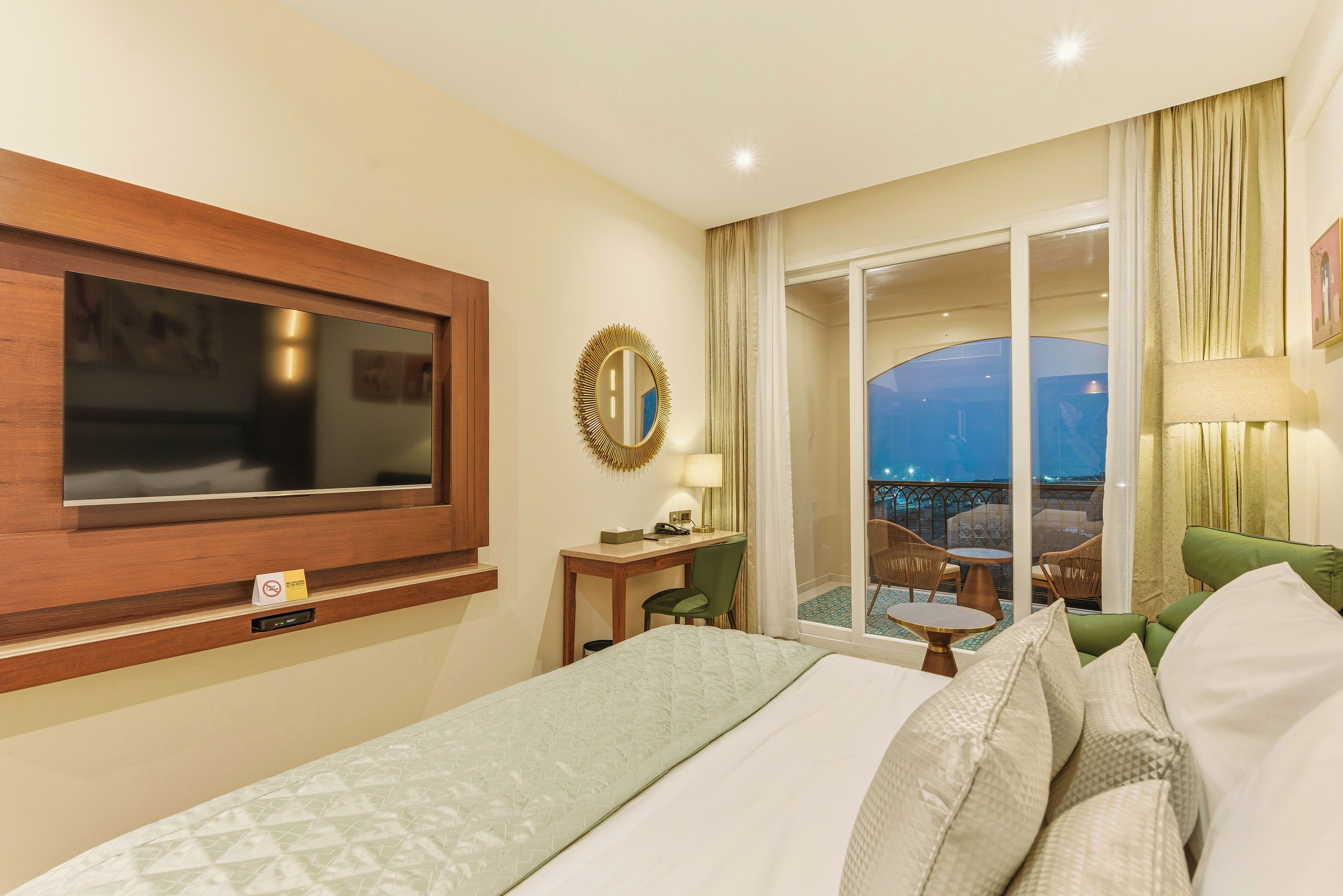 A hotel room with a bed, a TV mounted on the wall, a desk with a chair, a balcony with a table and chairs, and a view of the outside.