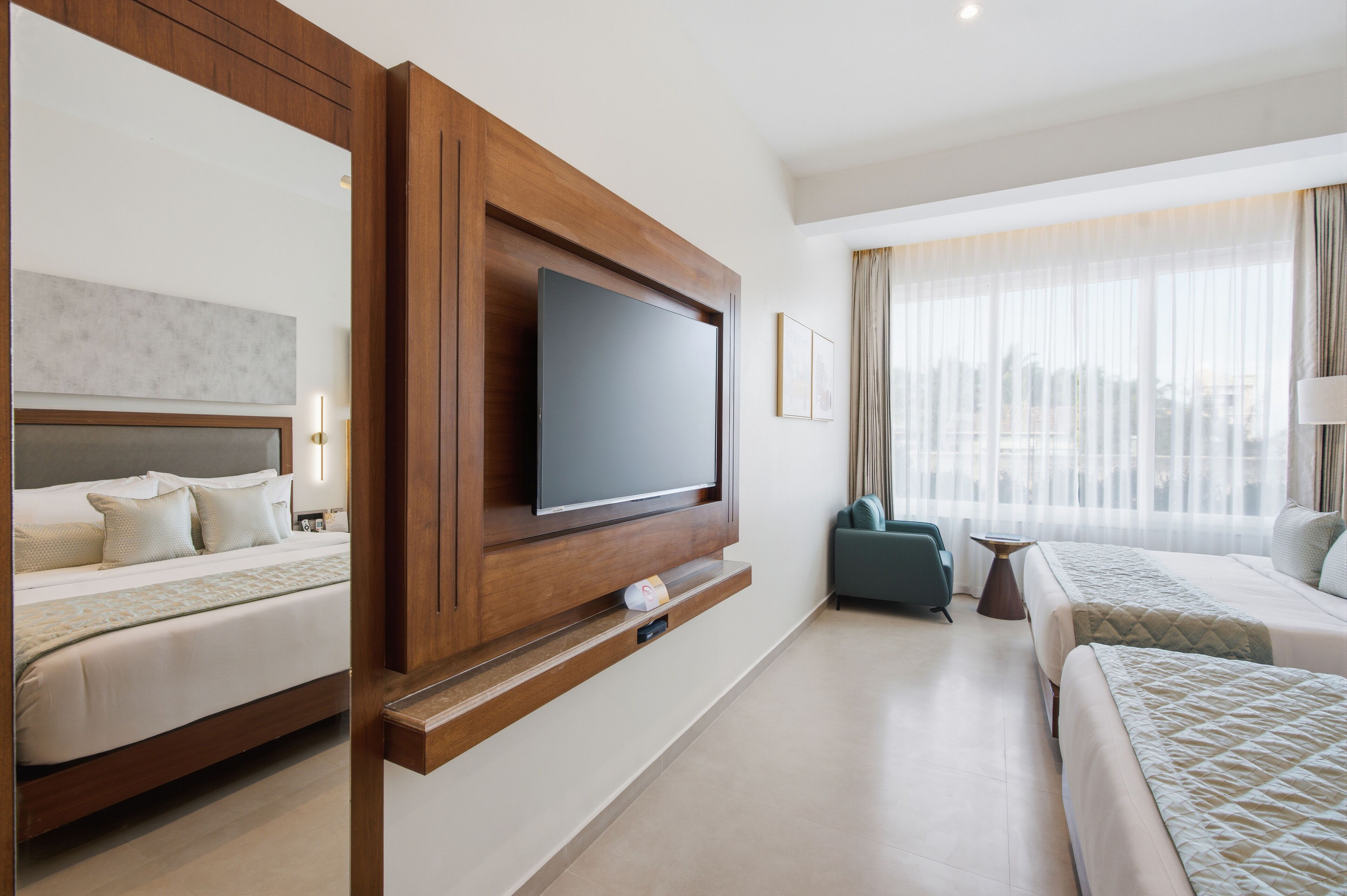 A hotel room with a large flat-screen TV mounted on a wooden panel, a bed with a headboard, a second bed, a small table, and a window with sheer curtains.