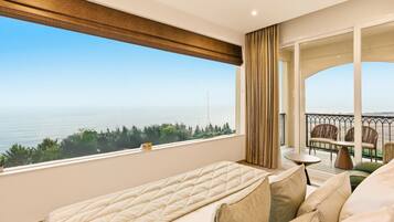 A hotel room with a large window offering a view of the sea and greenery. There is a bed, a small table, and a seating area with a view.