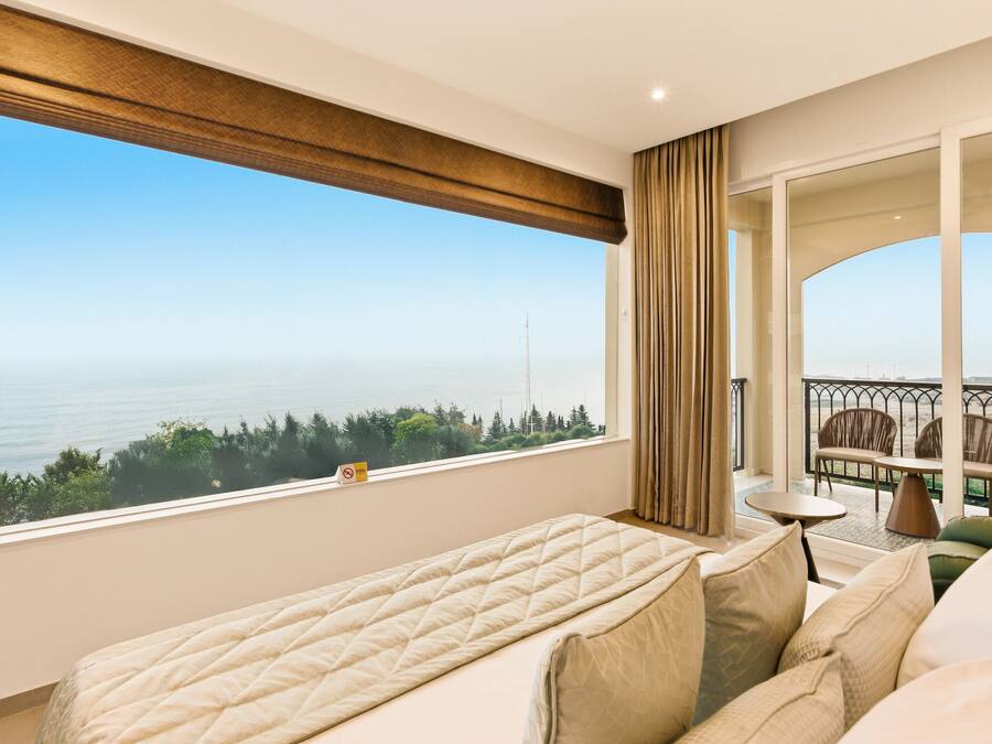 A hotel room with a large window offering a view of the sea and greenery. There is a bed, a small table, and a seating area with a view.
