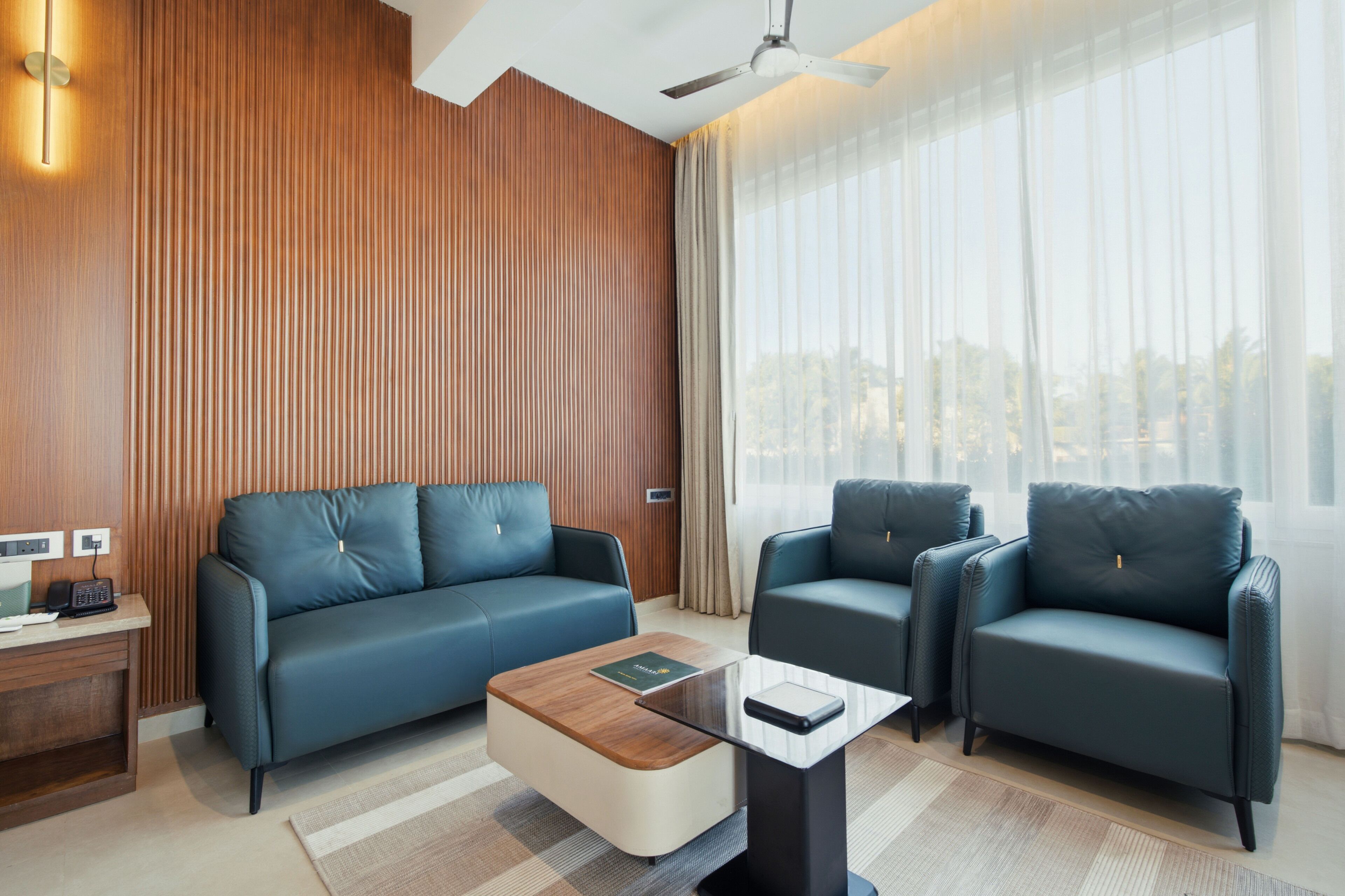 A modern hotel room with a wooden accent wall, a sofa, two armchairs, a coffee table, and a view through sheer curtains.