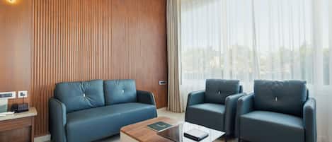 Une chambre d’hôtel moderne avec un mur d’accent en bois, un canapé, deux fauteuils, une table basse et une vue à travers des rideaux transparents.