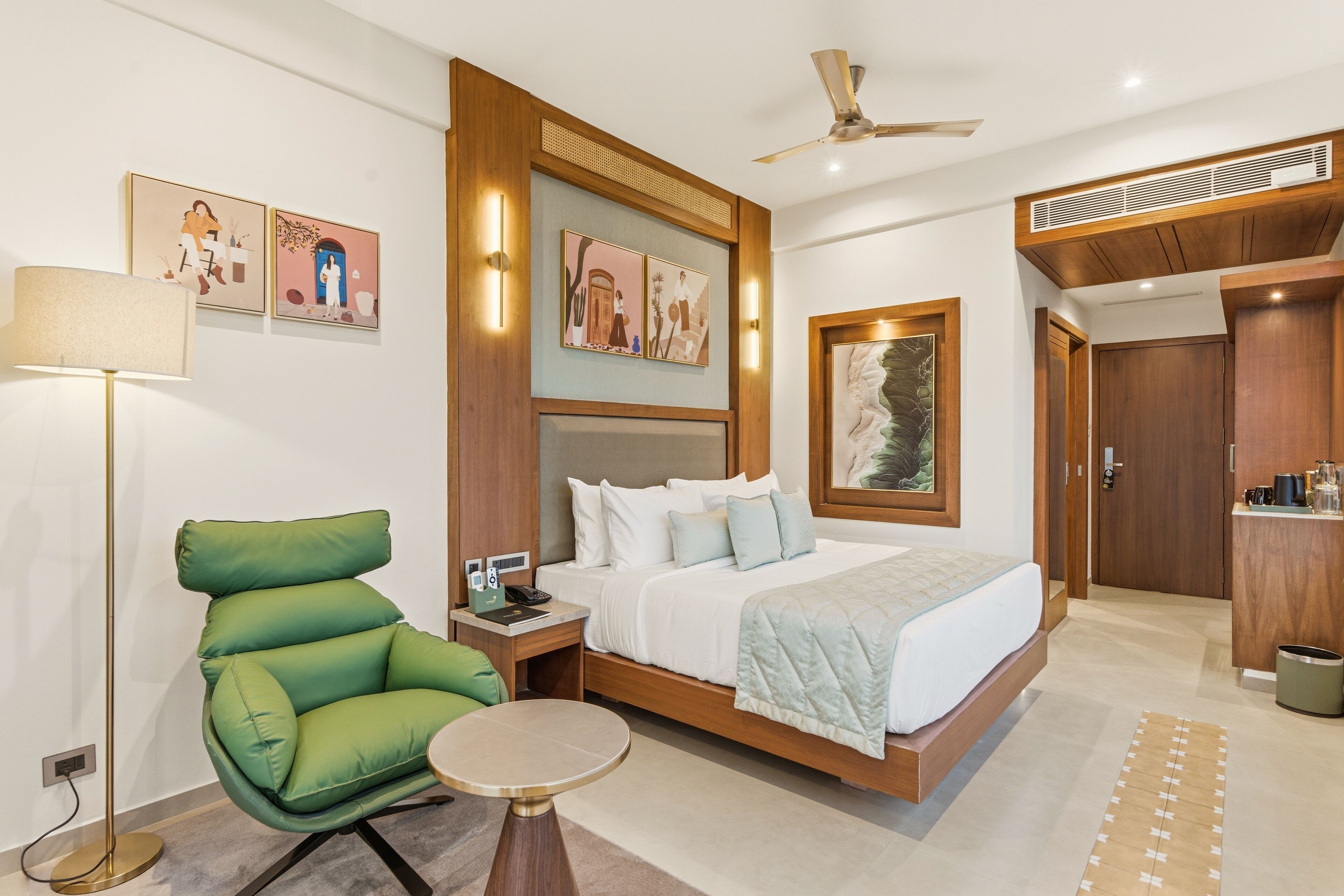 A modern hotel room with a bed, green chair, bedside table, lamp, artwork, and a kitchenette.