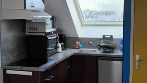 Cocina compacta con armarios oscuros, microondas, cafetera y lavamanos. Se ve un tragaluz sobre el lavamanos, y un bote de basura.