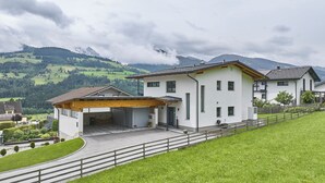 Una casa moderna de dos pisos con un porche cubierto, un camino de entrada pavimentado y una vista de colinas verdes y montañas al fondo.