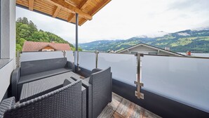 Un espacio al aire libre techado con mobiliario de mimbre, vista a un paisaje montañoso y un edificio con techo rojo al fondo.
