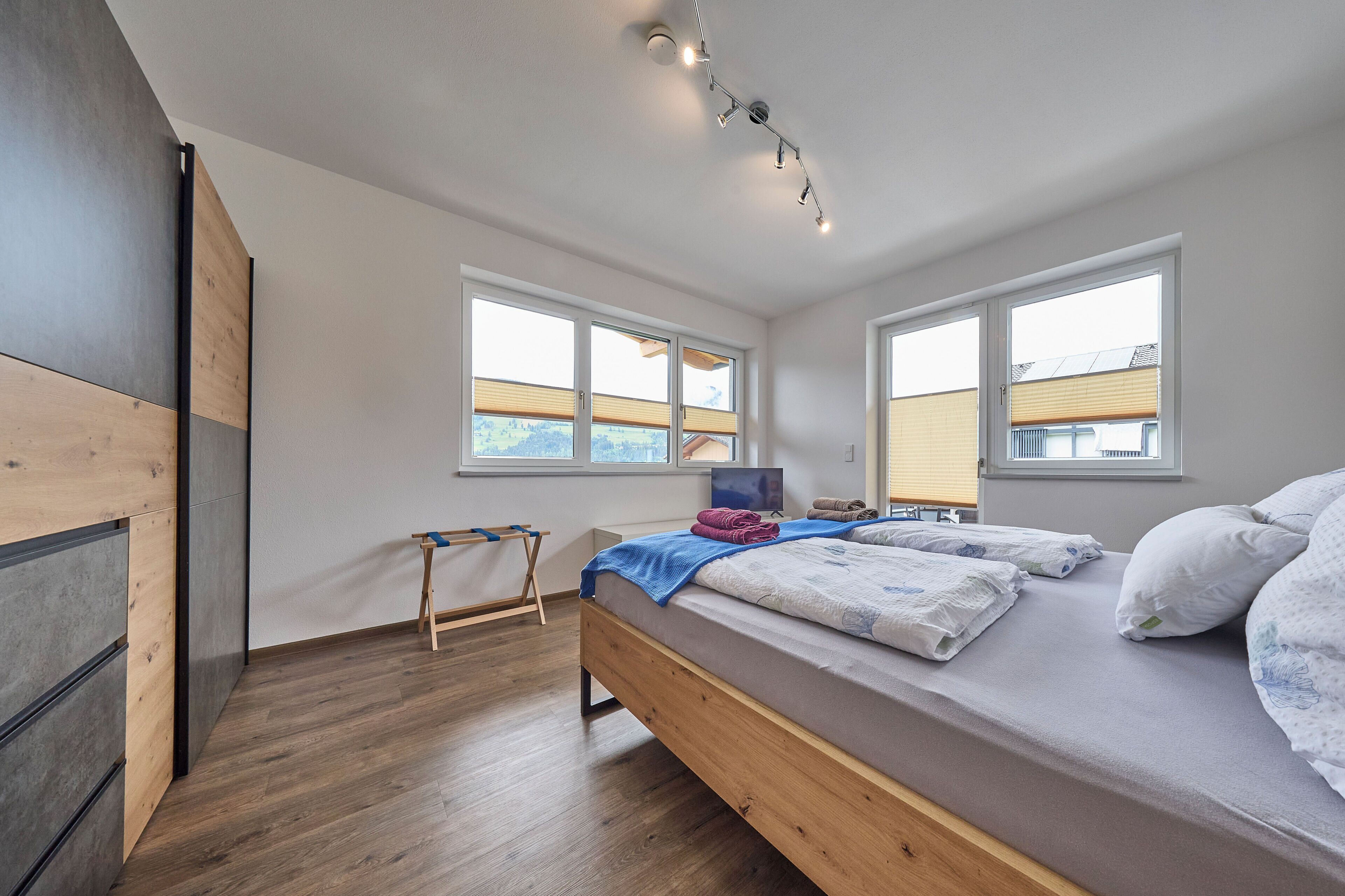 A modern bedroom with a wooden bed, two windows, a desk, and a chair.