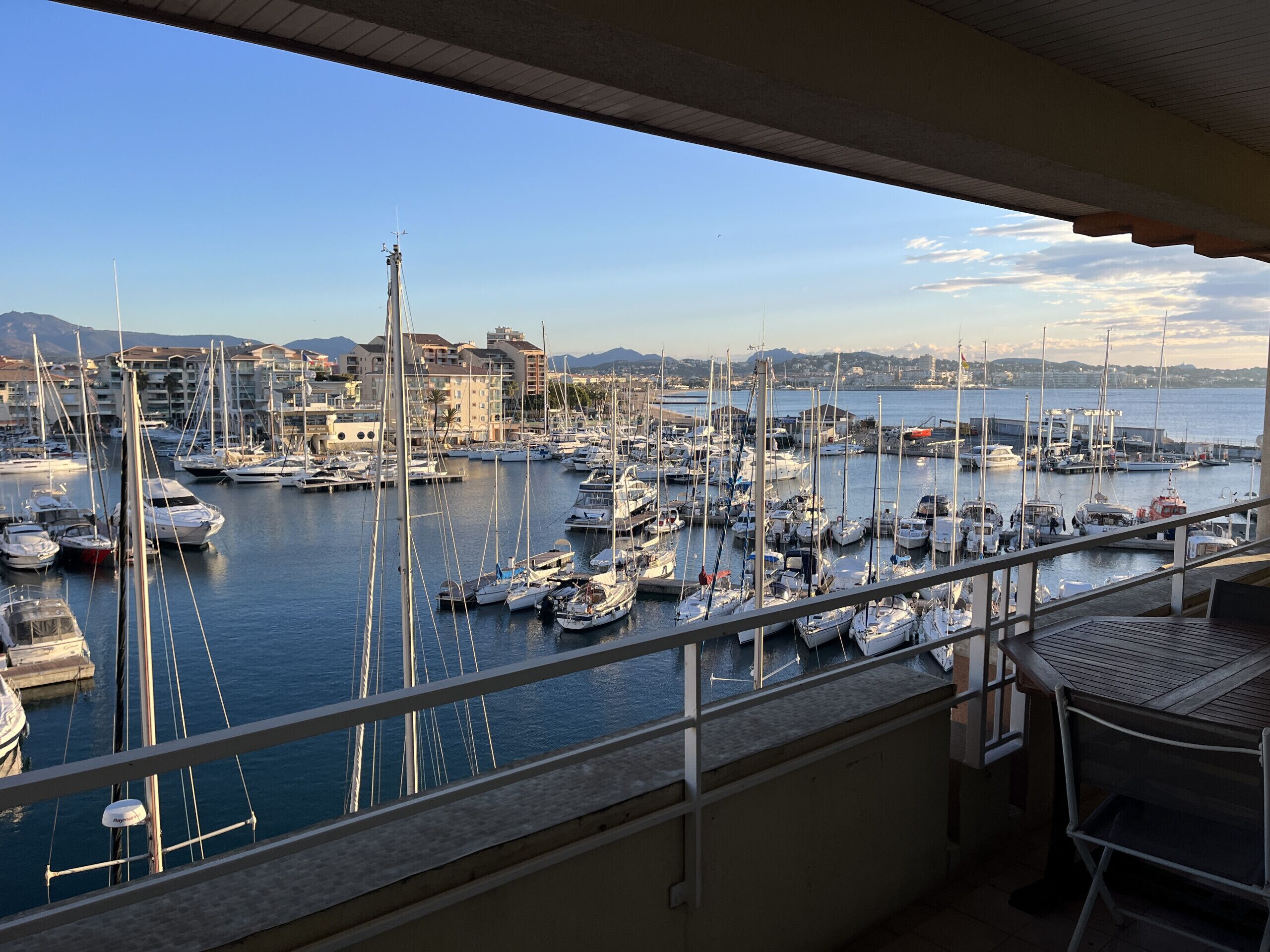 A marina with numerous boats docked, a balcony with a view of the marina, and buildings in the background.