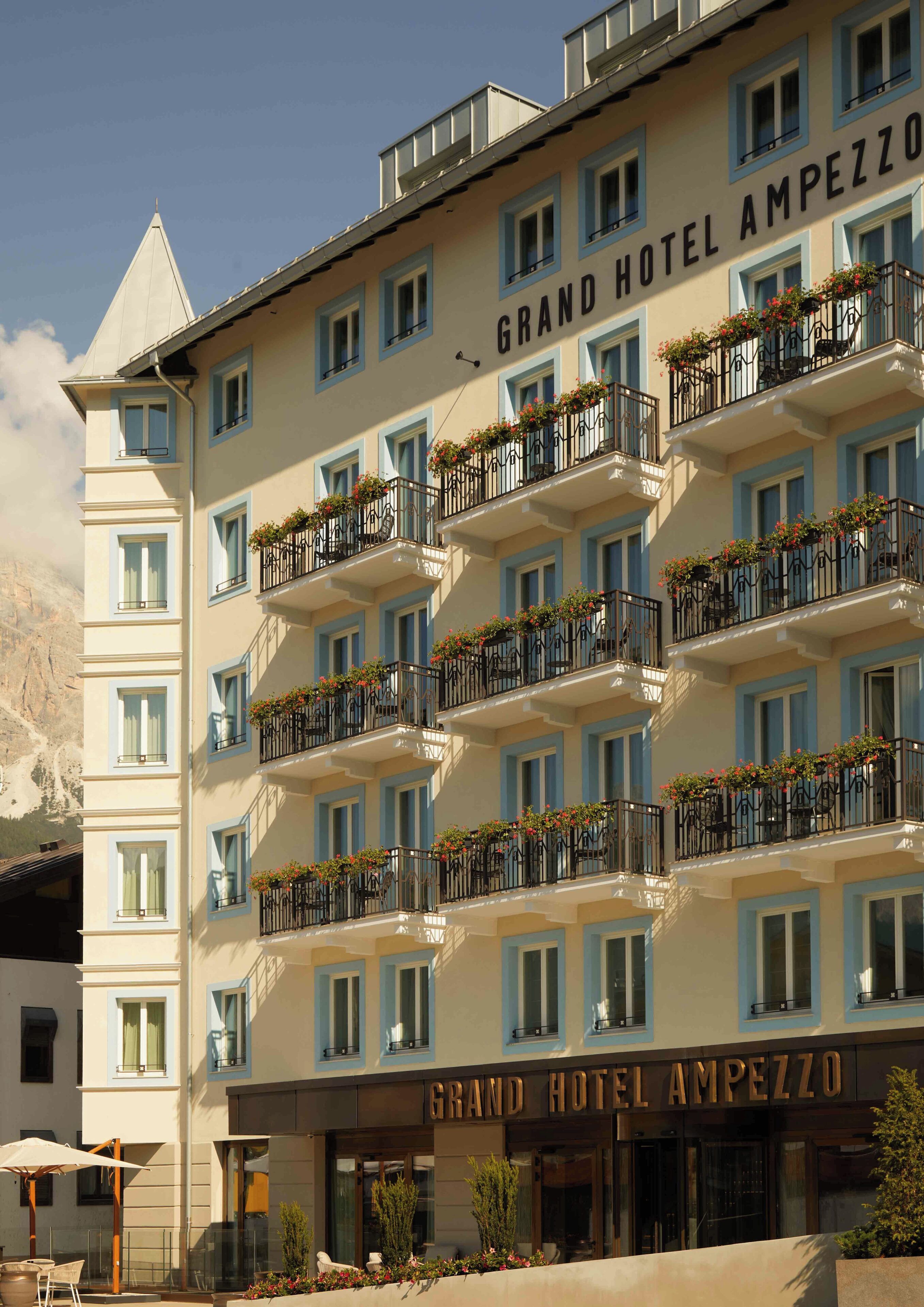A multi-story hotel with balconies, a sign reading 'Grand Hotel Ampezzo,' and a mountainous backdrop.