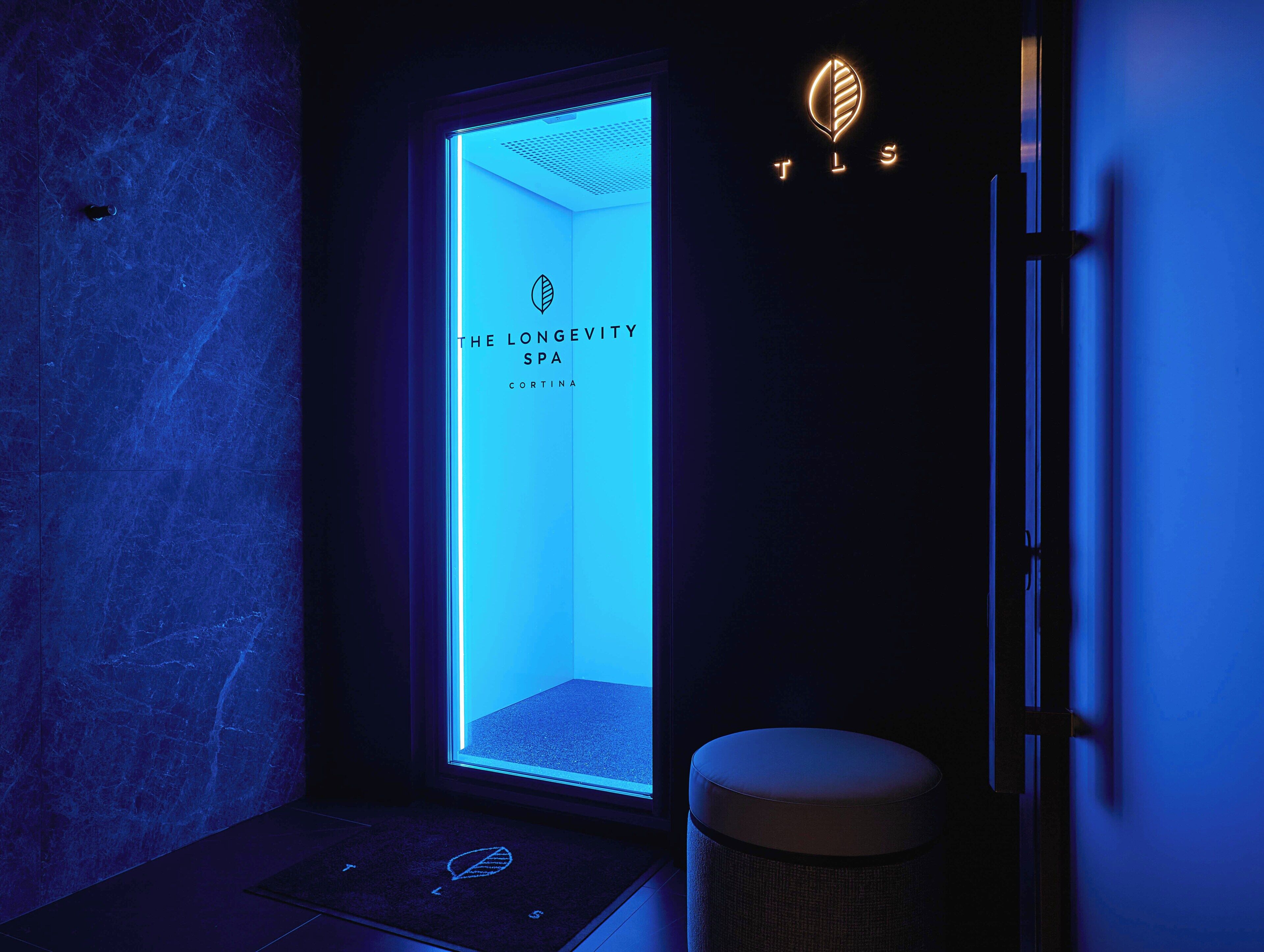 A spa room with a blue-lit door, a leaf motif on the door mat, and a wall-mounted sign for 'The Longevity Spa Cortina.'