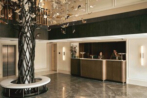 Un área de recepción con diseño moderno, que incluye un mostrador de recepción, un gran árbol decorativo y un piso de mármol.