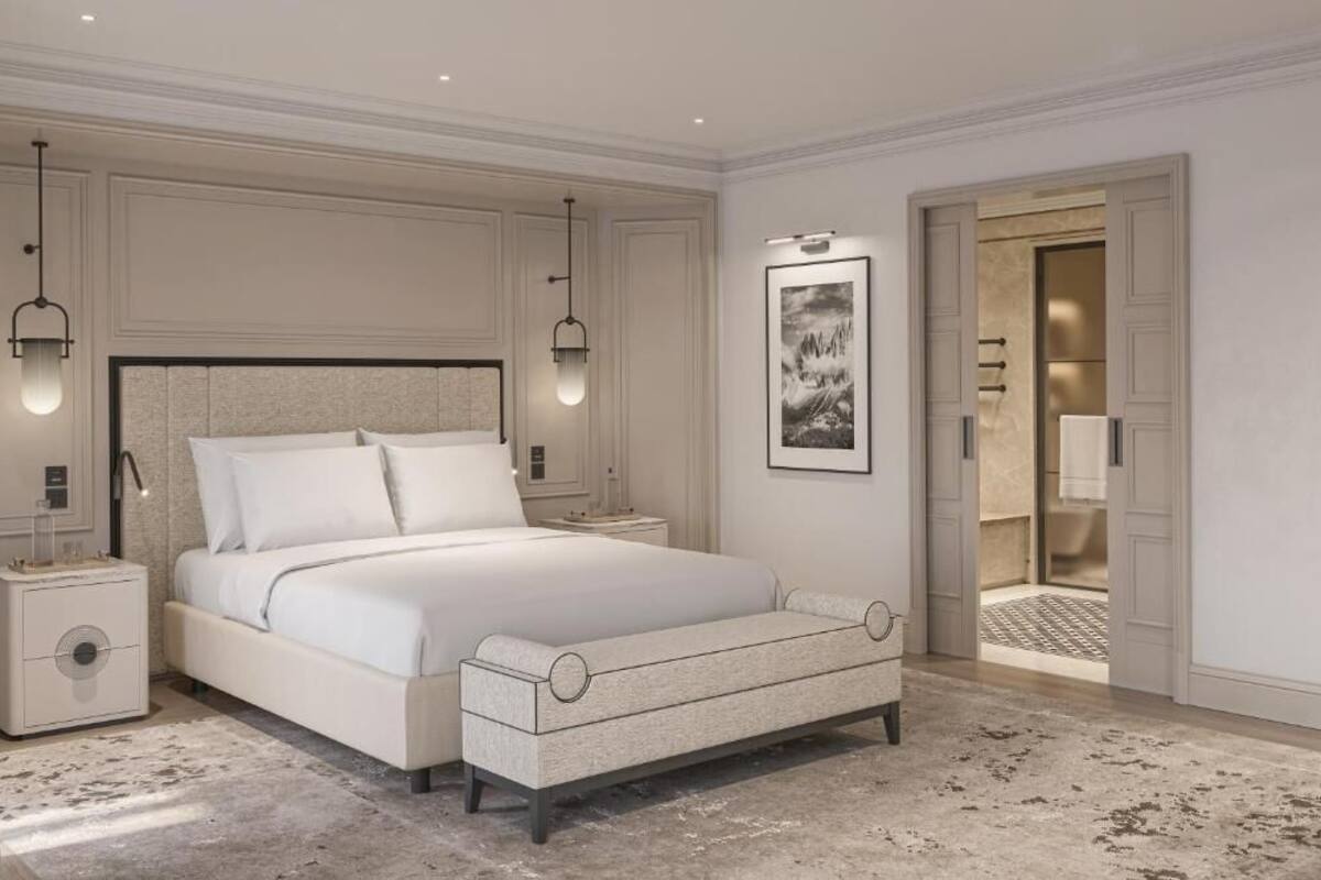 Un dormitorio con una cama grande, mesitas de noche, un banco, un cuadro enmarcado en la pared y una puerta abierta que da a otra habitación.