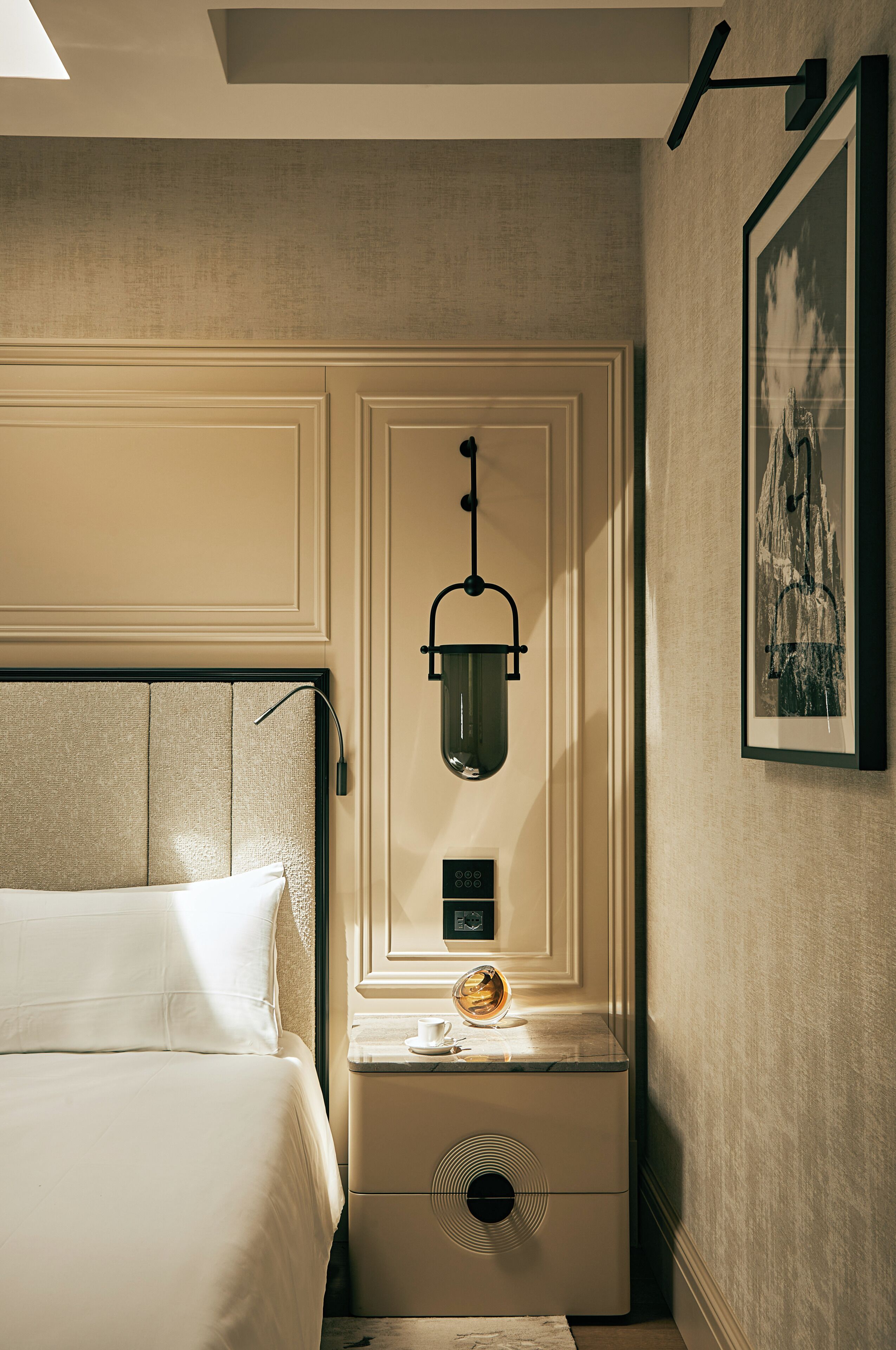 A hotel room with a bed, a bedside table with a lamp, a small table with a cup, and a framed picture on the wall.
