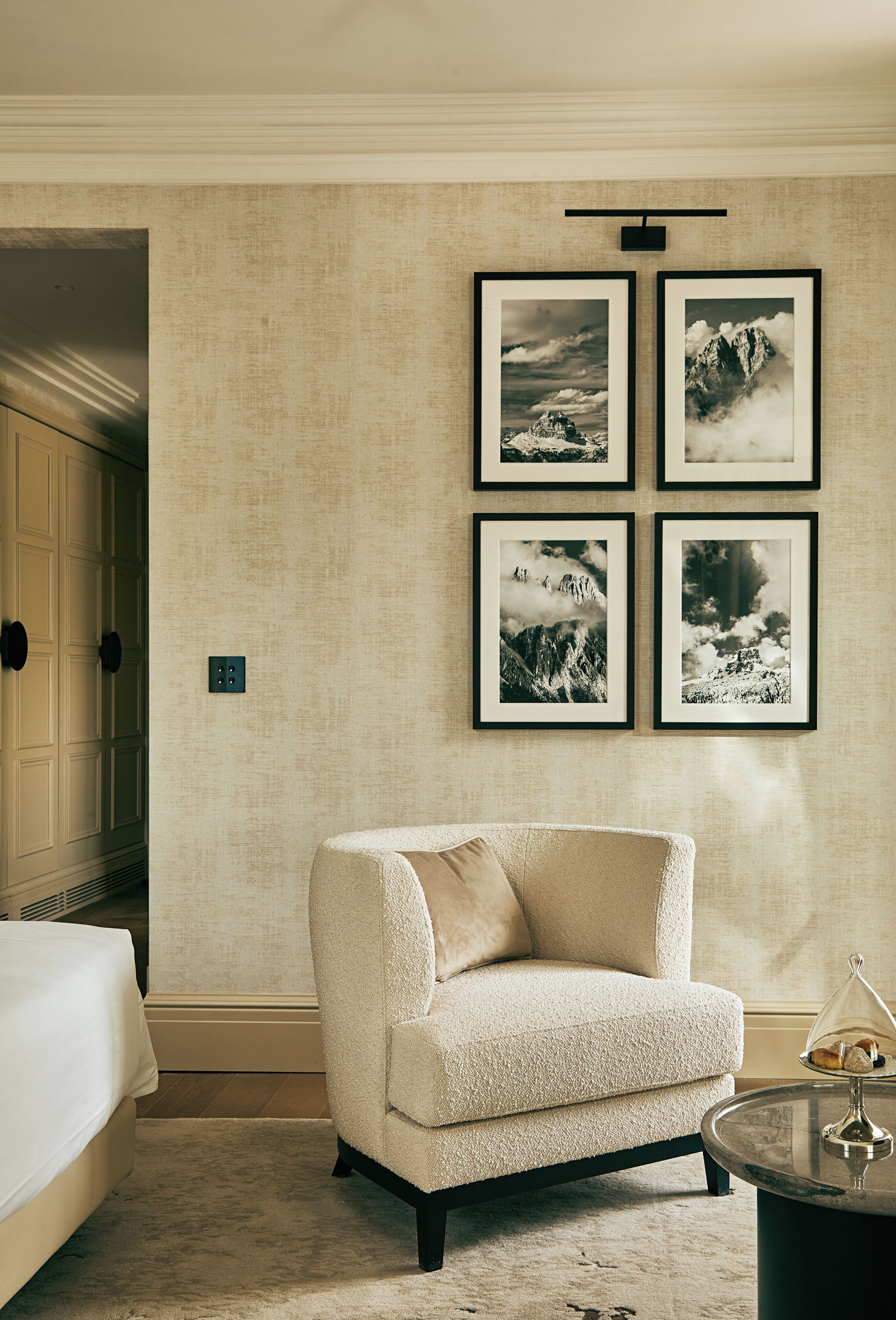 Habitación de hotel con pared beige con textura, un sillón blanco, una pequeña mesa redonda con una cúpula de vidrio y cuatro fotografías enmarcadas de paisajes montañosos.