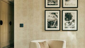 Habitación de hotel con pared beige con textura, un sillón blanco, una pequeña mesa redonda con una cúpula de vidrio y cuatro fotografías enmarcadas de paisajes montañosos.