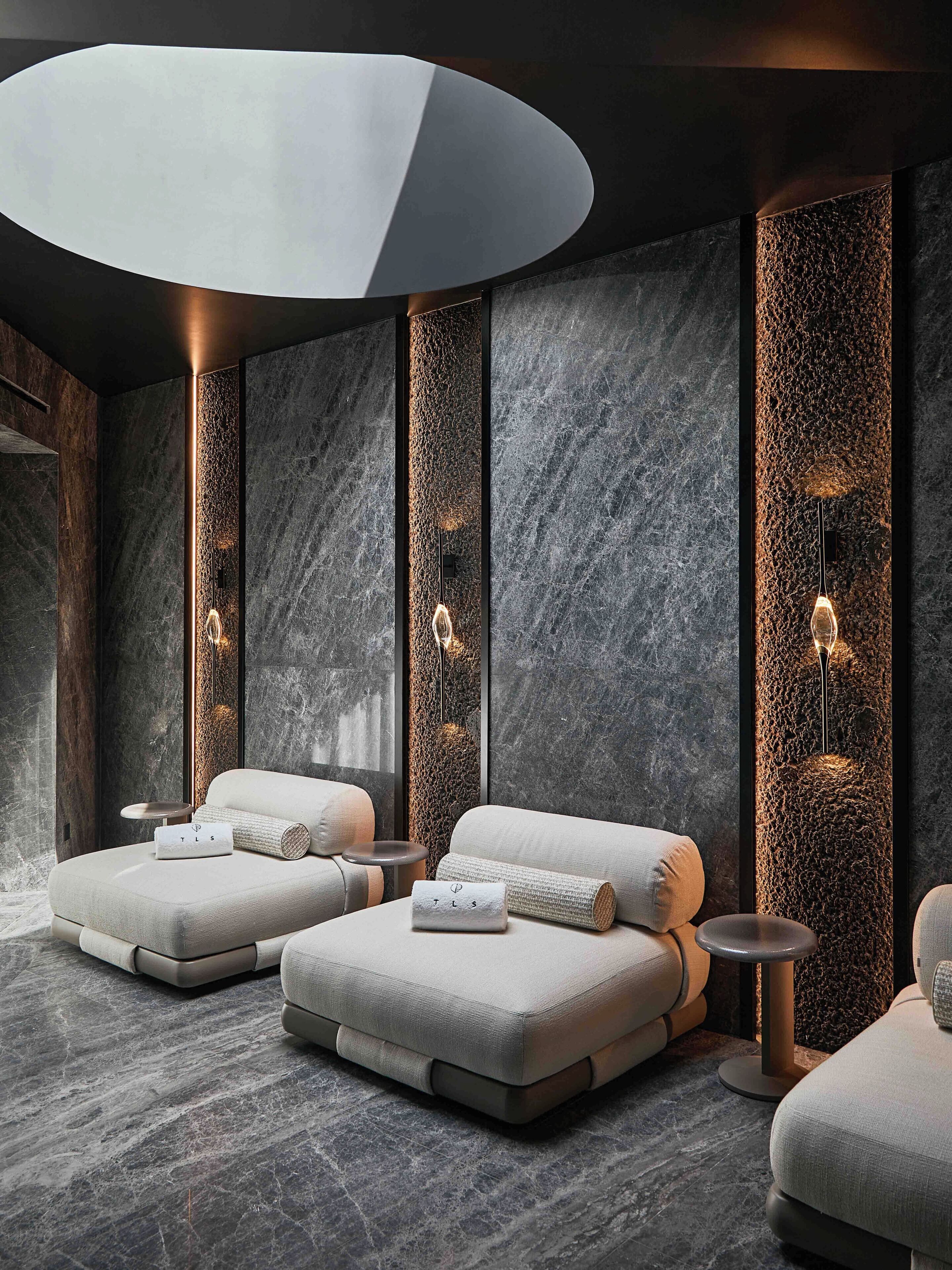 Ein moderner Wellnessraum mit zwei Sitzgelegenheiten, einem runden Tisch und einer wandmontierten Leuchte.