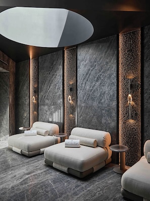 A modern spa room with two cushioned seating areas, a round table, and a wall-mounted light fixture. - Grand Hotel Ampezzo (Cortina d'Ampezzo)