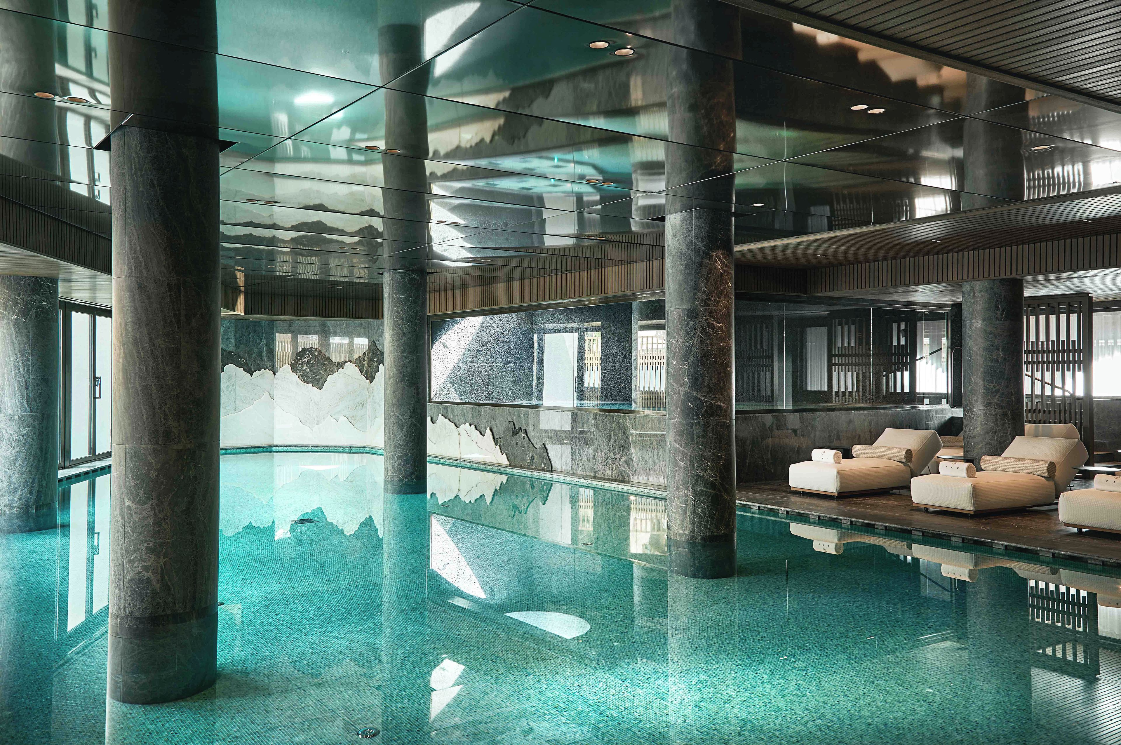 A modern indoor pool area with clear glass walls, large pillars, and lounge seating.