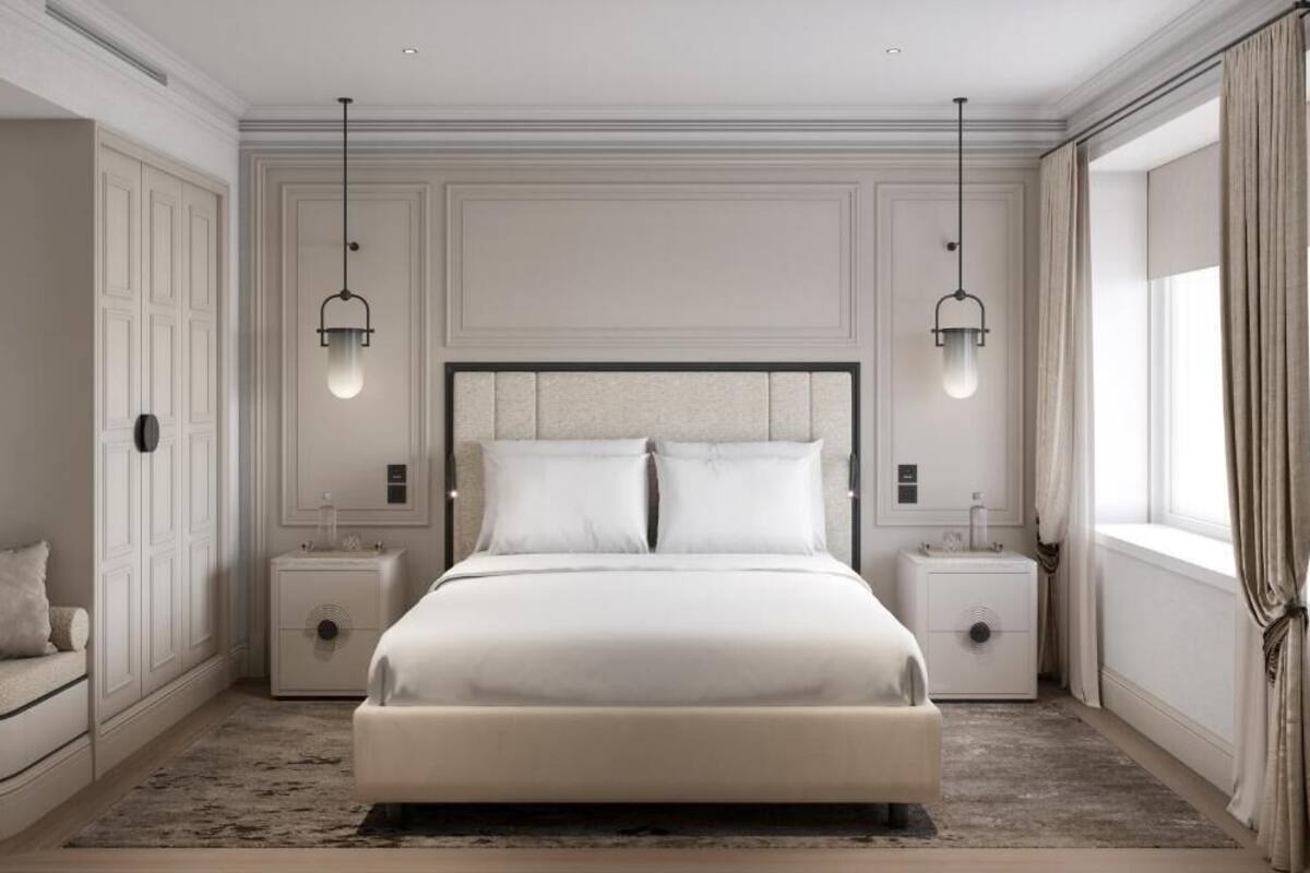 Un dormitorio con una cama grande, dos mesitas de noche, una lámpara auxiliar y una silla, además de un ventanal con cortinas.