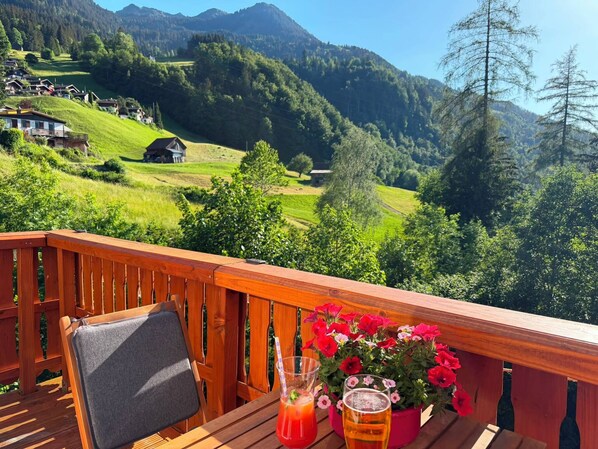 A wooden deck with a table, a chair, a glass of beer, and a pitcher of a red drink, overlooking a mountainous landscape with houses and greenery.