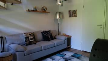 Een moderne woonkamer met een grijze bank, decoratieve kussens, een vloerlamp en een klein houten plankje met spullen.