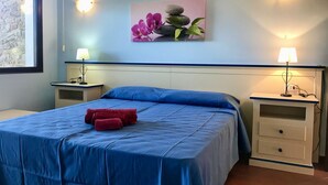 A bedroom with a blue bedspread, two bedside tables with lamps, a wall-mounted painting of flowers and stones, and a stone wall to the left.