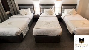 A hotel room with three beds, each with white bedding and a logo, a nightstand with a lamp, and artwork on the wall. - Petra Castle Hotel (Wadi Musa)