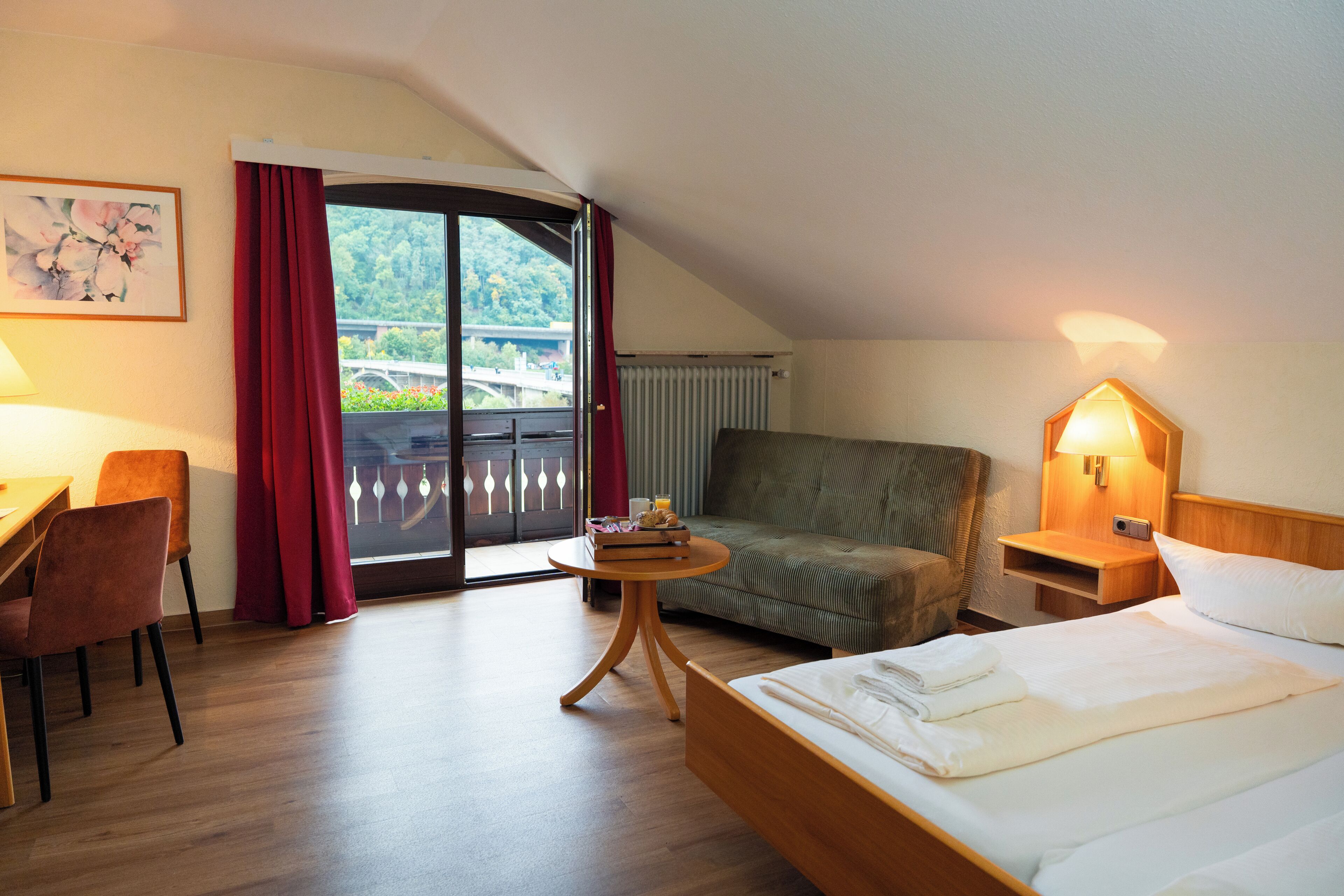 Ein Hotelzimmer mit Bett, Stuhl, Tisch und Blick auf eine Brücke und Grünanlage durch eine Schiebetür.