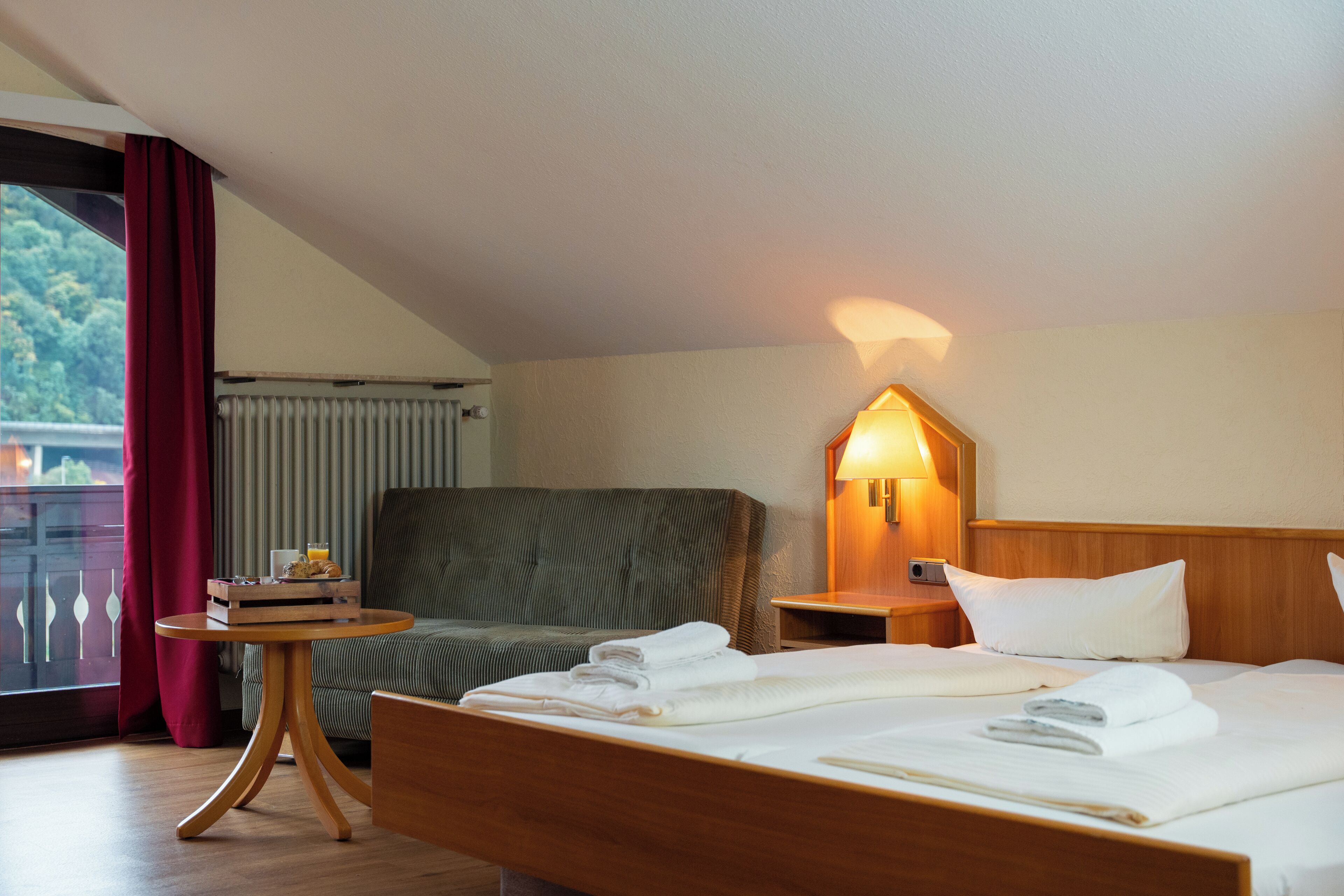 Habitación de hotel con una cama, una silla, una mesita con provisiones para el desayuno, una mesita de noche con una lámpara y vistas a zonas verdes.