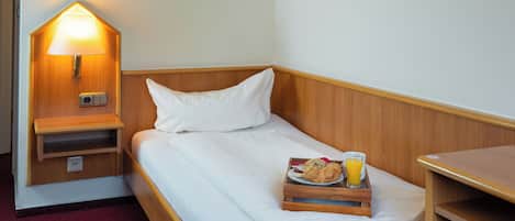 A single bed with a wooden headboard, a bedside table with a lamp, a tray with food and a drink, and a framed picture on the wall.