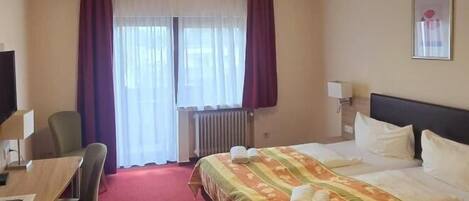 A hotel room with a bed, a desk, a chair, a radiator, and a window with curtains.