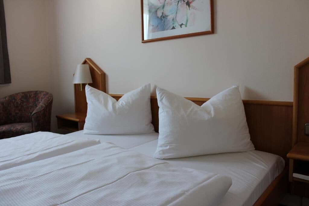 A hotel room with a large bed, two pillows, a nightstand with a lamp, a chair, and a painting on the wall.