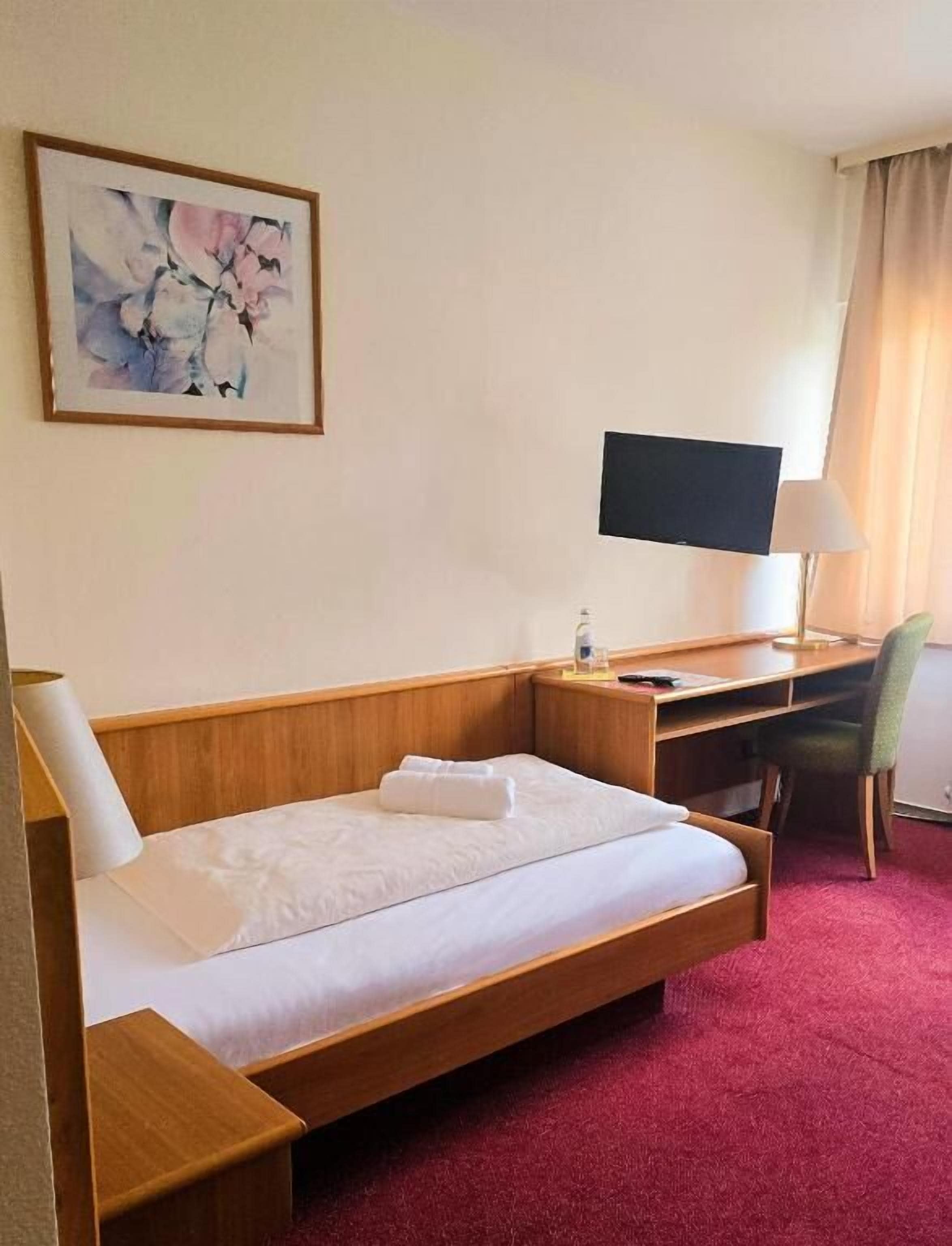 A hotel room with a bed, a desk, a chair, a TV, and a painting on the wall.