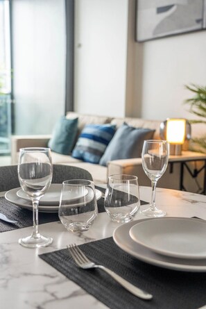 A dining table set with plates, glasses, and cutlery, with a sofa and lamp in the background. - The Starcatcher’s Nest - Lv 33 Skyhome on The Star (Brisbane City)