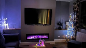 Ein modernes Wohnzimmer mit einem an der Wand befestigten Flachbildfernseher, einem Kamin mit farbigem Licht und einem Weihnachtsbaum.
