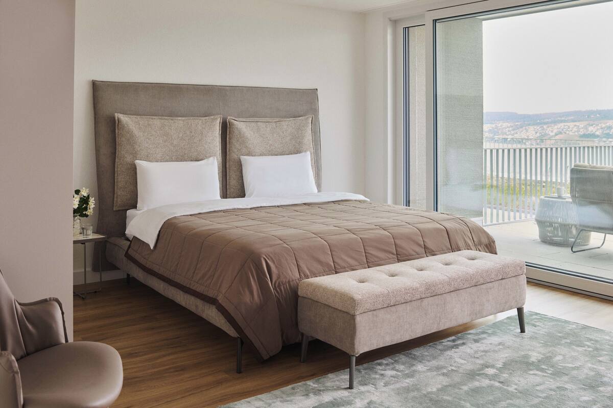Un dormitorio moderno con una cama grande, un banco, una mesita de noche y vistas al exterior.