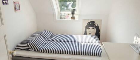 Une petite chambre à coucher, bien rangée, avec un lit simple, une fenêtre et une étagère fixée au mur, ornée d’objets décoratifs.