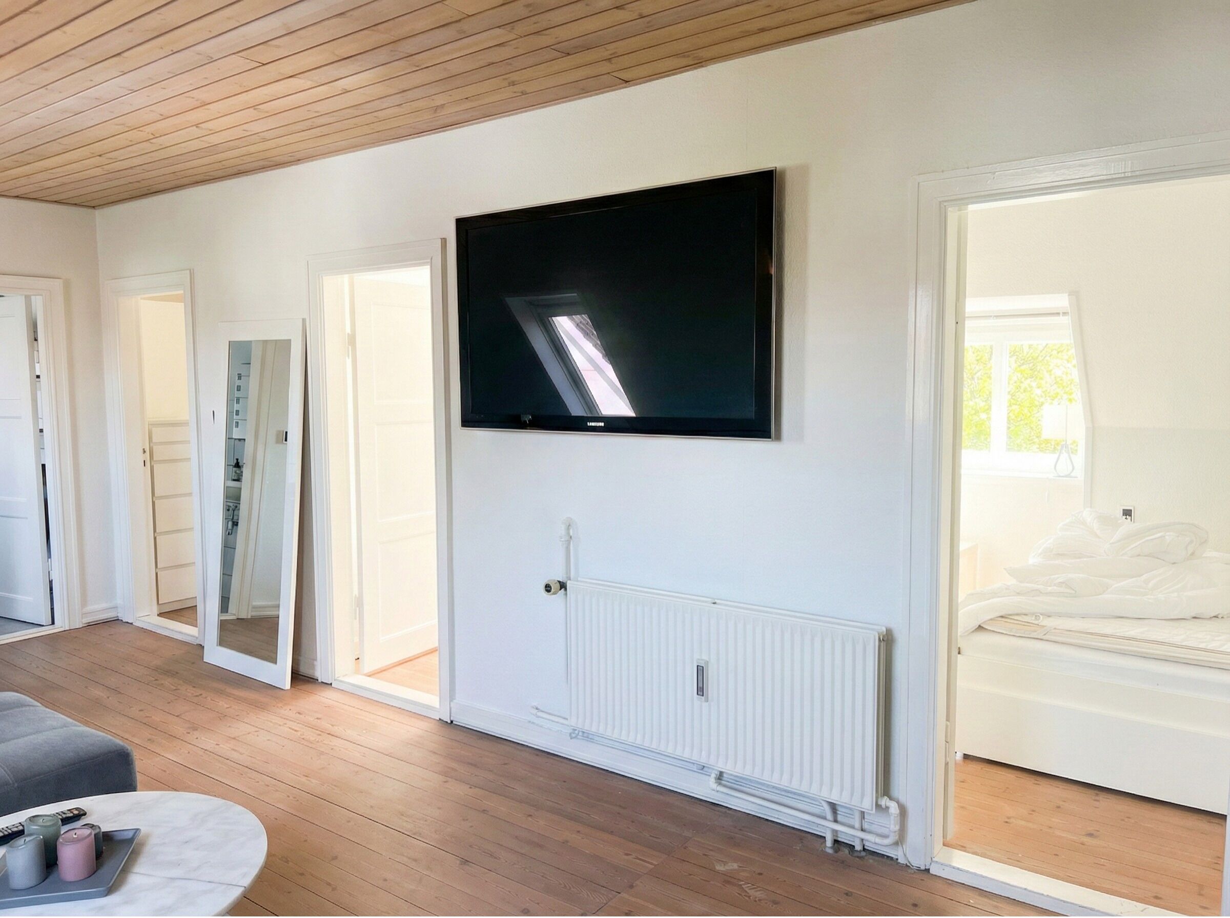 A room with a flat-screen TV mounted on the wall, a radiator below it, and a bed with white bedding to the right.