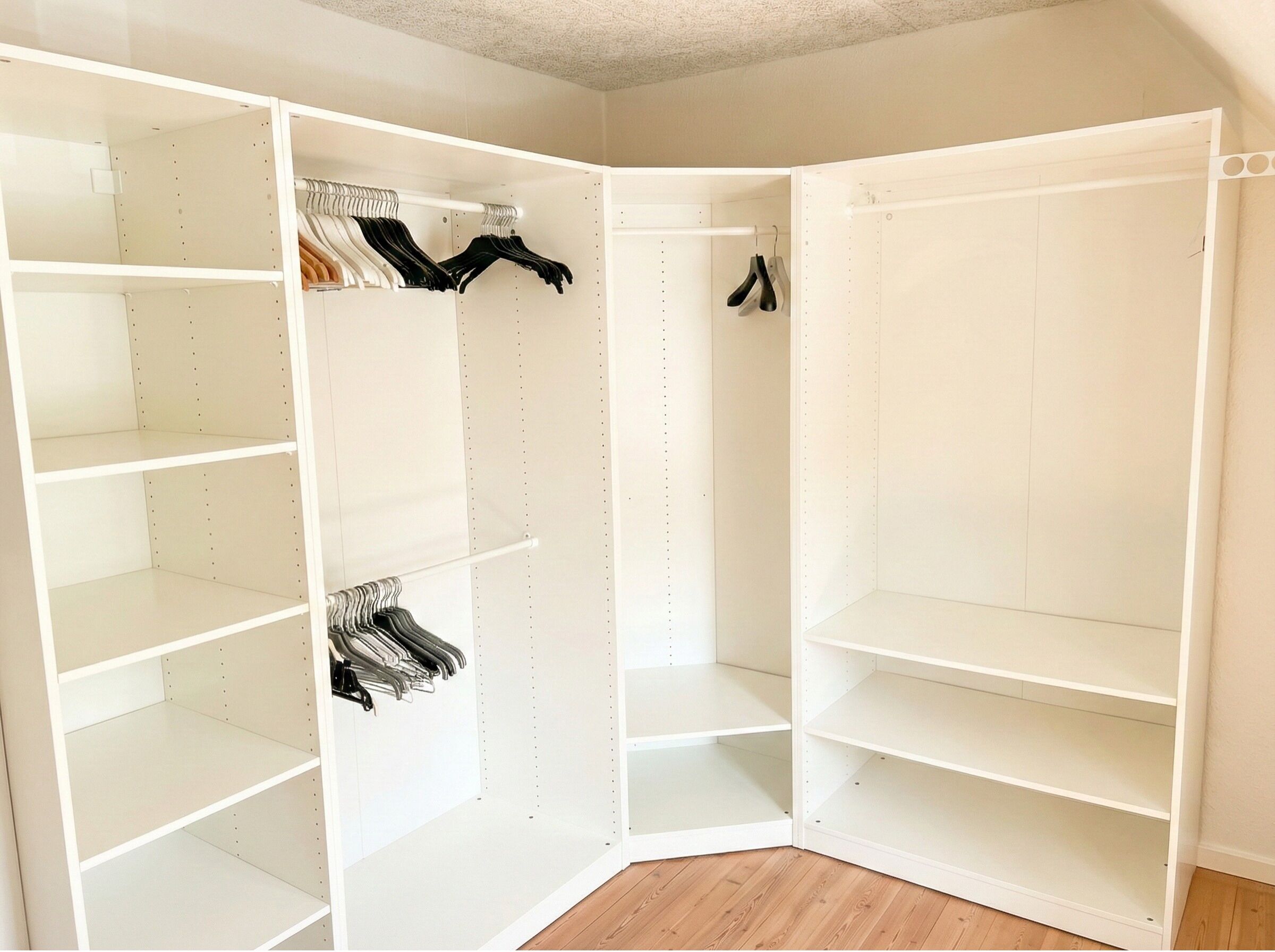 A corner closet with multiple shelves and hanging space, featuring a white interior and wooden flooring.