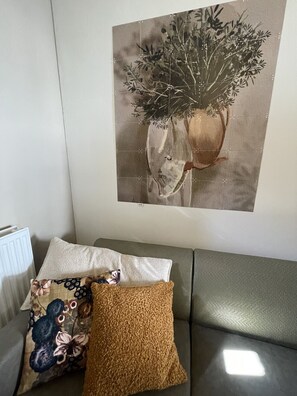 A living room with a grey sofa, a brown textured pillow, and a floral pillow. A framed artwork of a plant in a vase is on the wall. - 18. 4p Authentic Cosy Cottage (Voerendaal)